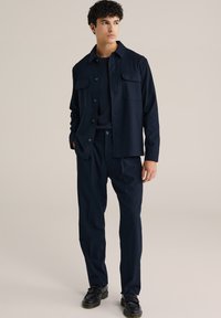 Marinblå avslappnad outfit med en knäppjacka med två bröstfickor, matchande byxor och svarta skor med detaljerad textur.