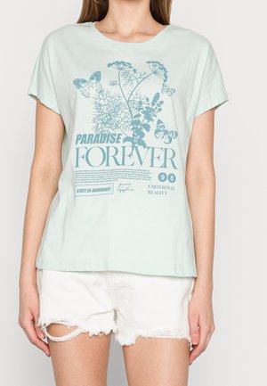 Camiseta de algodón verde menta claro con mangas cortas, que presenta un estampado botánico y texto en azul más oscuro. Lleva puesta con pantalones cortos blancos deshilachados.