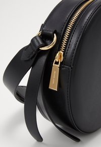 Sac à main rond en cuir noir avec fermeture éclair dorée et anneau pour sangle, avec tirette de fermeture éclair dorée à logo.