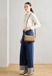 Vävd beige crossbody-väska med bruna läderaccenter, tillsammans med en ljus långärmad skjorta och vida denimjeans med frans.