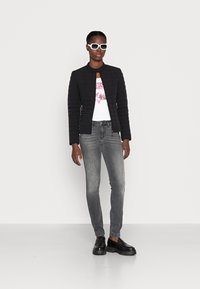 Zwarte gewatteerde jas, wit graphic t-shirt, grijze skinny jeans en zwarte chunky instappers. Model draagt witte zonnebril. Effen achtergrond.
