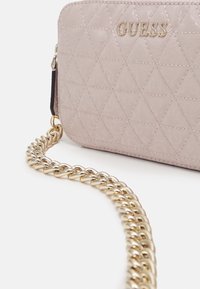 Prošívaná růžová syntetická kabelka se zlatým řetízkovým popruhem. Na přední části má embosovaný vzor a logo značky. Kompaktní a strukturovaný design.