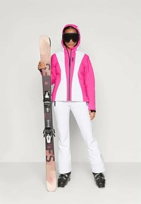 Smučarska oprema z živo roza in belo jakno, belimi hlačami in črnimi smučarskimi čevlji. Drži smuči s vezmi in roza robovi.