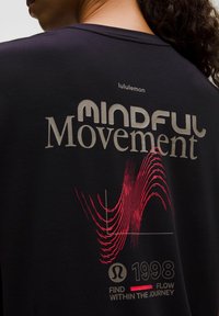 Czarna koszulka z krótkim rękawem z grafiką "Mindful Movement" w metalicznej srebrnej i czerwonej falistej linii, z logo lululemon powyżej i rokiem 1998 poniżej.