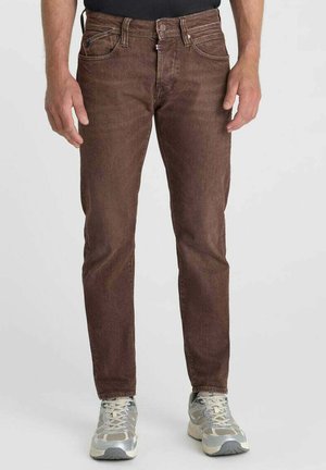 Uomo con jeans marroni slim fit e scarpe da corsa grigie e bianche in piedi davanti a uno sfondo semplice.
