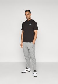 Čierne tričko s logom na hrudi, v kombinácii so svetlosivými teplákmi a bielymi športovými topánkami. Hladká tkanina a voľný strih.