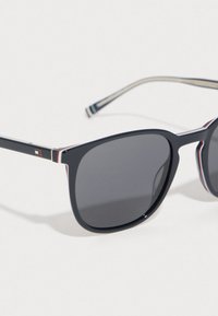 Čierne slnečné okuliare so tmavými sklami, plastový rám so subtílnymi červenými a bielymi pruhmi pozdĺž ramena a detail loga na boku.