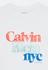 Bílý bavlněný tričko s barevným, vrstveným textem: "Calvin" v červené, "Klein" v světle modré a "nyc" v modré. Obsahuje značkovou etiketu uvnitř.
