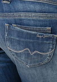 Jeans de mezclilla azul con un bolsillo trasero, con costuras plateadas y un patrón de olas, marcados con "Pepe Jeans" en cursiva.