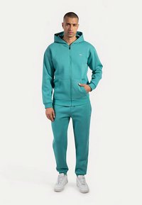 Turkooizen zip-up hoodie met een voorzak en bijpassende broek. Gemaakt van zachte stof, voorzien van een capuchon en minimale merkdetails. Inclusief witte sneakers.