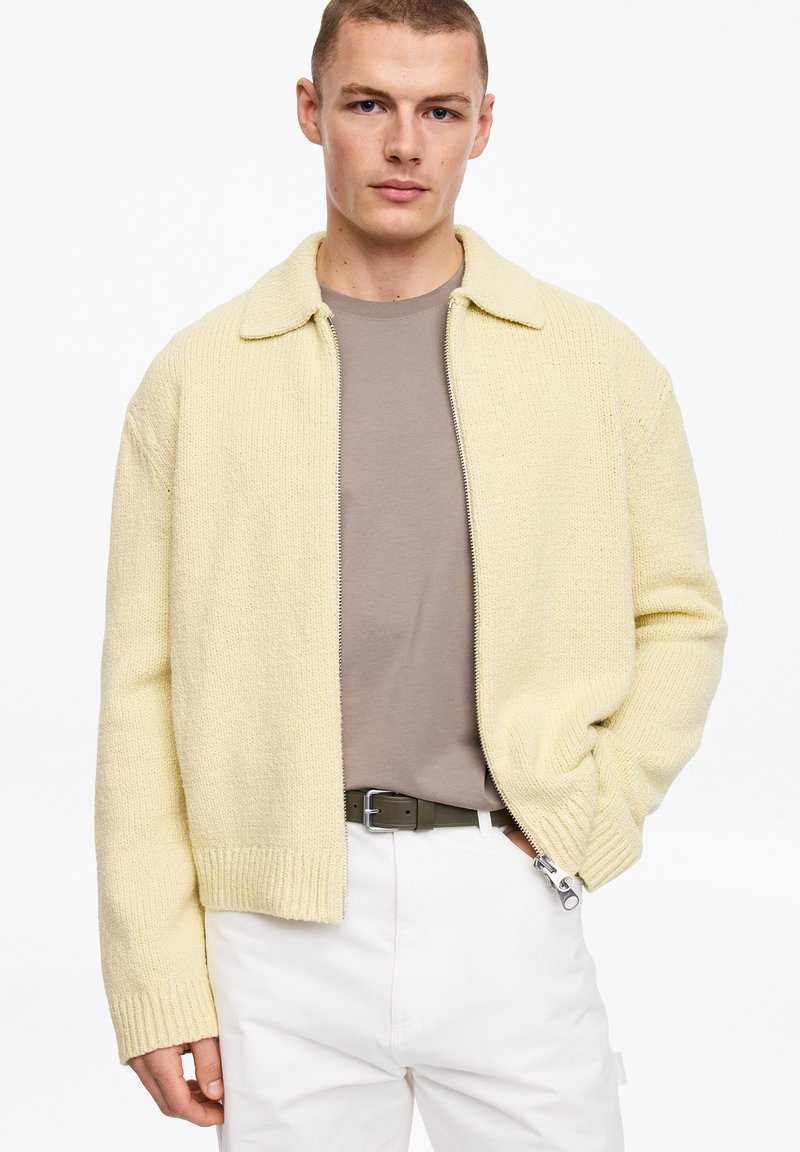 Jeune homme portant un cardigan zippé texturé jaune pâle sur un t-shirt beige, un pantalon blanc et une ceinture foncée, debout avec une main dans la poche.