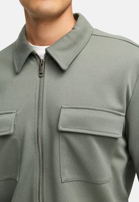 Olivgrüne Zip-Jacke aus strukturiertem Stoff, mit einem Kragen, zwei Brusttaschen und einer weißen Innenschicht.