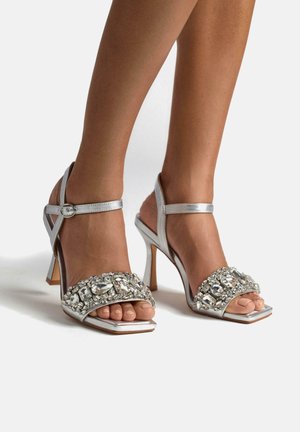 Sandalias de tacón alto plateadas con punta cuadrada y una detallada correa joya, con correas en el tobillo y un acabado brillante y elegante.