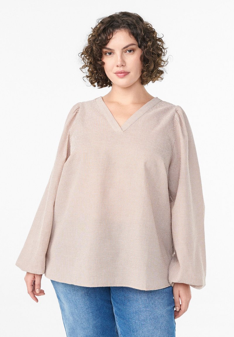 Svetloružová, štruktúrovaná blúzka s výstrihom do V a zvonovými dlhými rukávmi. Je vo voľnom strihu a má bodkovaný vzor, kombinovaná s modrými džínsami.