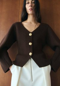 Frau trägt eine dunkelbraune V-Ausschnitt-Strickjacke mit drei goldenen Knöpfen und weiße, hochtaillierte Hose mit vorderem Reißverschluss, steht vor einer Holzvertäfelung.