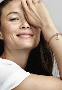 Bracciale dorato con un ciondolo a forma di cuore che presenta una pietra rosa. Modello con lunghi capelli e pelle imbellita da lentiggini, sorride su uno sfondo neutro.