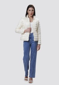Giacca piumino bianca con zip sul davanti e colletto alto, indossata sopra una maglietta bianca e jeans a vita alta azzurri. Texture liscia, design minimale.