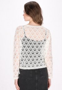 Cardigan in pizzo color crema con un motivo a rombo, texture semi-trasparente e maniche lunghe, indossato sopra una maglia nera. Il bordo presenta un rifinitura scallop.