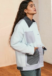 Lichtblauwe jas met grijze corduroy kraag, opgestikte zak en contrasterende grijze accenten. Bevat voorzakken en een knoopsluiting.