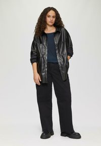 Schwarze Kunstlederhemdjacke mit großen Taschen, kombiniert mit einem dunkelblauen Oberteil und schwarzen Weitbein-Hosen. Schwarze Stiefel mit dicker Sohle.