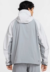 Chaqueta ligera con capucha en gris y blanco de corte relajado. Incluye mangas largas y un logo negro de Nike cerca del dobladillo.