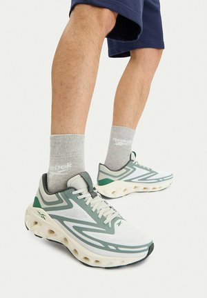 Nogi w szarych skarpetkach Reebok i zielono-białych sportowych butach Reebok z masywnymi podeszwami, stojące na jasnym tle.