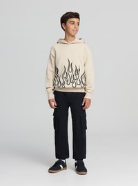Beżowa bluza z kapturem z czarnym wzorem płomieni u dołu, noszona z czarnymi spodniami typu cargo i czarnymi trampek, stojąca na neutralnym tle.