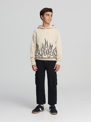 Felpa con cappuccio beige con motivo a fiamma nera all'orlo, indossata con pantaloni cargo neri e scarpe da ginnastica nere, in piedi davanti a uno sfondo neutro.