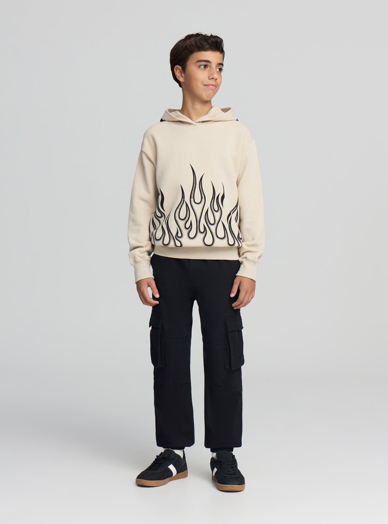 Beżowa bluza z kapturem z czarnym wzorem płomieni u dołu, noszona z czarnymi spodniami typu cargo i czarnymi trampek, stojąca na neutralnym tle.