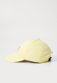 Gul bomullslue med en buet skygge, med en orange logo foran, justerbar stropp bak, og synlige sømdetaljer.