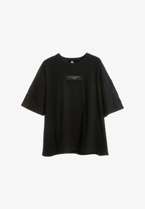 Camiseta negra oversize con una pequeña etiqueta rectangular en el pecho y un bordado negro parecido a plumas en ambas mangas.