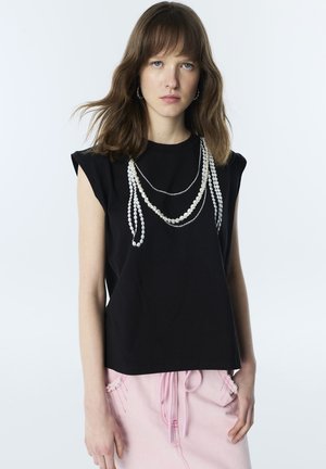 Mujer con cabello castaño hasta los hombros viste una camiseta negra sin mangas, collares de perlas en capas y pantalones rosas con cordón.