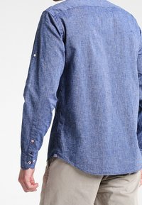 Mand iført en langærmet blå struktureret skjorte og beige bukser, vist fra ryggen mod en ensfarvet hvid baggrund.