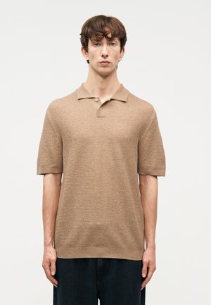 Fiatal férfi rövid barna hajjal, bézs színű, texturált rövid ujjú galléros ingben és sötét nadrágban, sima háttér előtt állva.