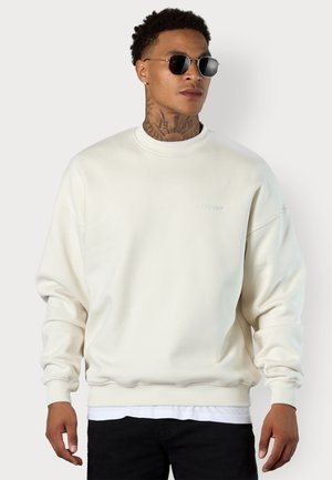 Cream-colored sweatshirt with a round neck, dropped shoulders, and ribbed cuffs. Features a subtle embossed logo on the chest.