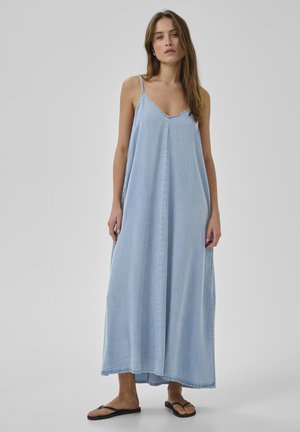 Nainen seisoo yllään löysä vaaleansininen hihaton maksimekko ja mustat varvastossut, neutraali tausta.