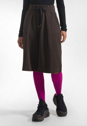 Persona con una falda marrón hasta la rodilla, calcetines altos de color rosa brillante, zapatillas negras y una camiseta negra de manga larga, de pie sobre un fondo blanco.
