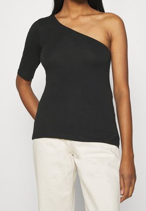 Femme portant un top noir asymétrique avec une manche courte et un pantalon blanc, debout devant un fond uni.