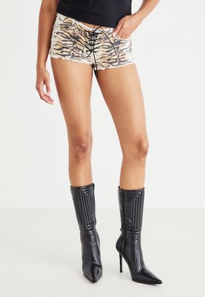 Mujer con pantalones cortos de encaje con estampado de tigre y botas negras acolchadas de tacón alto, de pie con una mano en el bolsillo contra un fondo blanco.