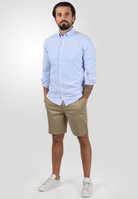 Camisa abotonada azul claro de mangas largas, pantalones cortos color caqui y zapatillas blancas. Modelo de pie con las manos en los bolsillos, fondo neutro.