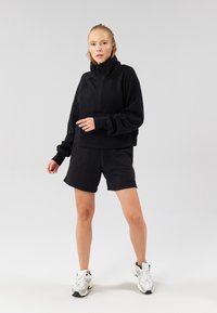 Mustast lühendatud pusast kõrge krae ja ees oleva taskuga, kombineeritud mustade lühikestega. Modell kannab valgeid tossusid, esitades lõõgastunud sportliku stiili.