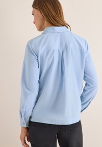 Light blue long-sleeve shirt with a collar, featuring a back pleat and buttoned cuffs. Soft fabric texture, styled for a relaxed fit.