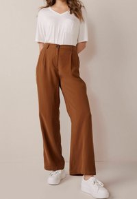 Braune, weit geschnittene Hose mit hoher Taille, ausgestattet mit Falten und einem Knopfverschluss. Kombiniert mit einem lockeren weißen V-Ausschnitt-T-Shirt und weißen Turnschuhen.