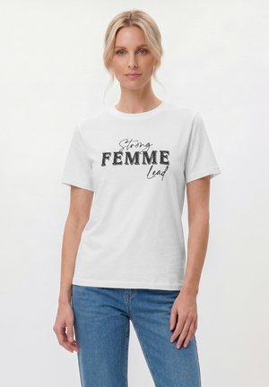 Ξανθιά γυναίκα που φοράει λευκό t-shirt με το κείμενο «Strong FEMME Lead» και μπλε τζιν, στέκεται μπροστά σε απλό λευκό φόντο.