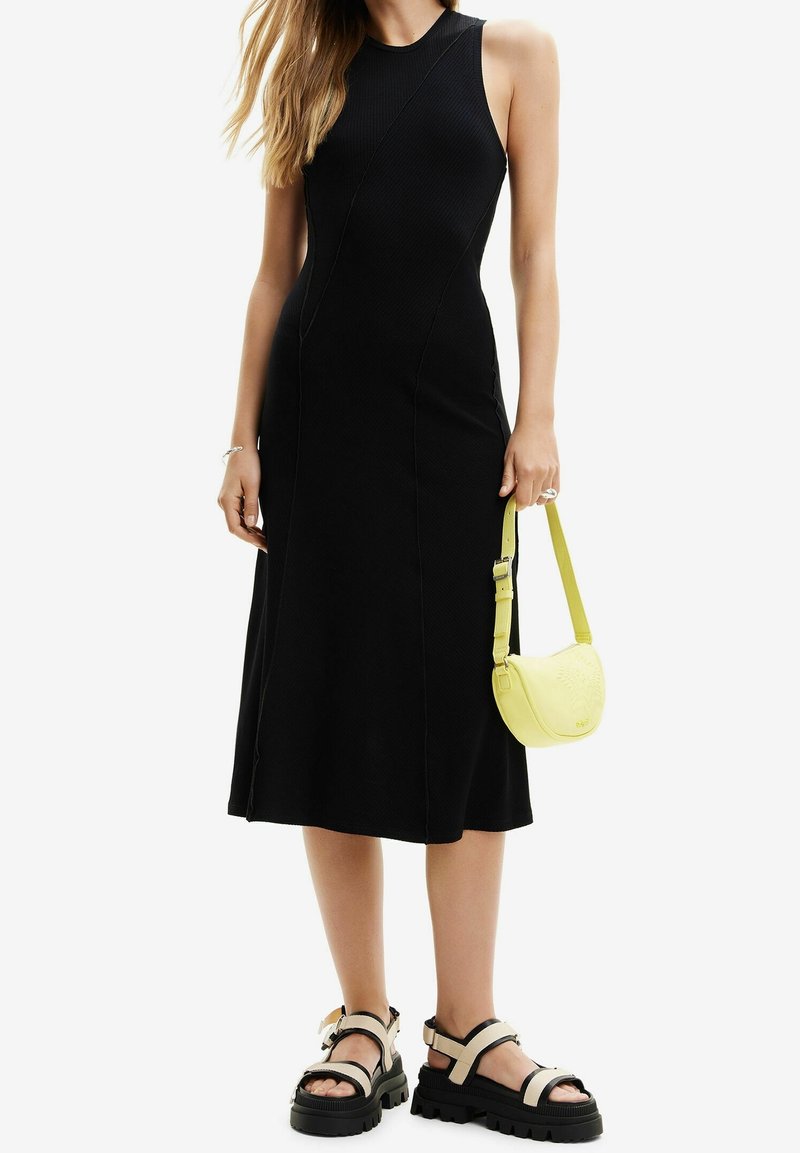 Musta ribbineulottu midimekko, jossa on vartalonmyötäinen siluetti, yhdistettynä paksupohjaisiin mustiin sandaaleihin ja pieneen keltaiseen olkalaukkuun.