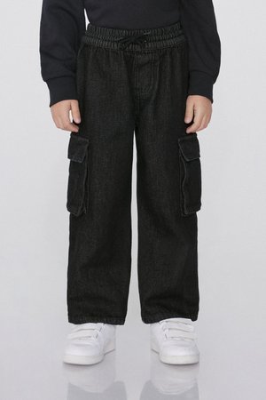 Dítě v černých cargo kalhotách s bočními kapsami a bílých teniskách na suchý zip, stojící před jednobarevným pozadím.