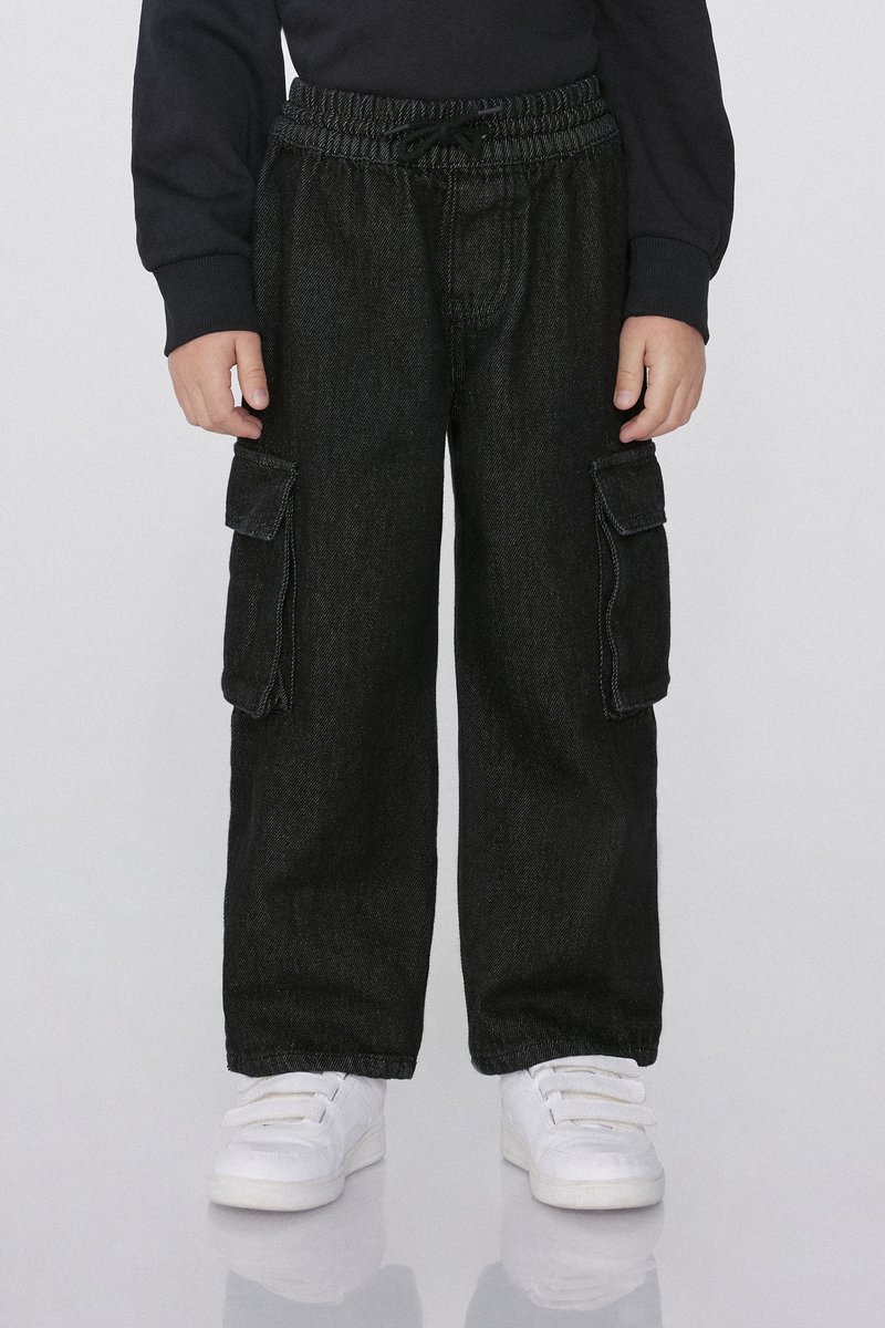 Kind trägt schwarze Cargo-Hose mit Seitentaschen und weiße Klettverschluss-Sneaker, steht vor einem einfarbigen Hintergrund.