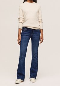 Maglione beige chiaro con polsini e orlo a coste, abbinato a jeans svasati blu scuro. Calzature sono scarpe da ginnastica avorio con suola testurizzata.