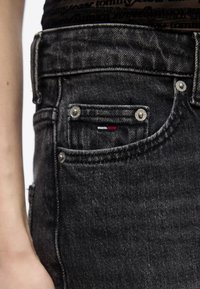 Κοντινό πλάνο ενός ατόμου που φοράει μαύρο τζιν, εμφανίζοντας την εμπρόσθια μικρή τσέπη με ένα ορθογώνιο λογότυπο σε κόκκινο, άσπρο και σκούρο μπλε χρώμα.