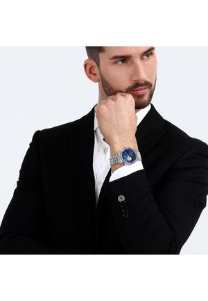 Silverfärgad armbandsur med en blå urtavla, band i rostfritt stål, bärs på handleden av en man i svart kostym och vit skjorta, handen positionerad eftertänksamt.